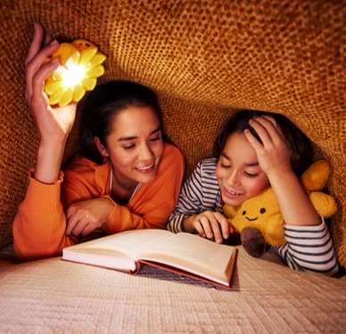 Zwei Personen lesen gemeinsam unter einer Decke, beleuchtet von einer kleinen Lampe, mit einem aufgeschlagenen Buch und einem Stofftier